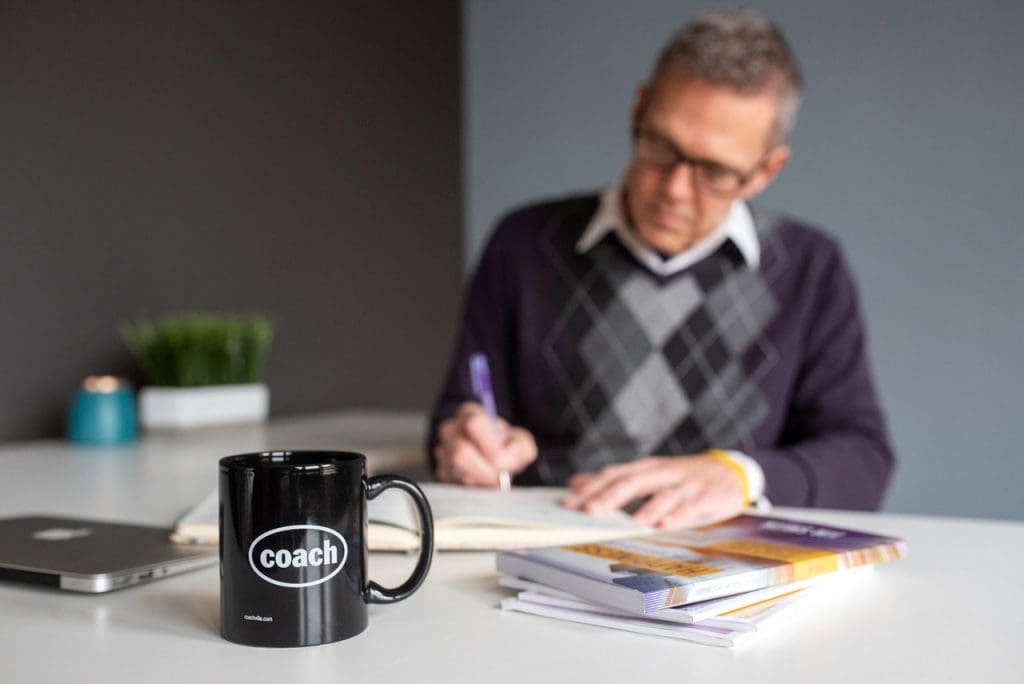 Act Happy Week, Day 4 - Jim sitting at a desk writing happiness exercises with a coffee cup that reads "Coach" in the forefront. 